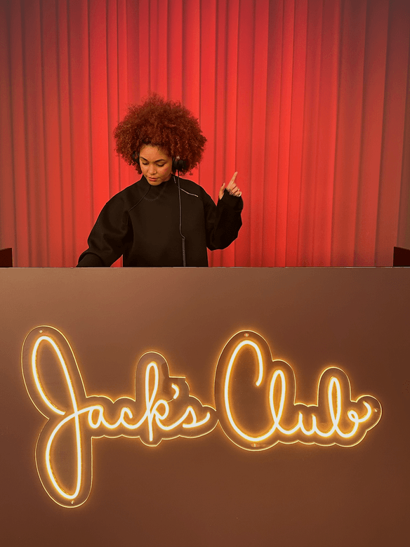 DJ de cabello rizado ante una cortina roja, detrás de un letrero de neón de Jack's Club
