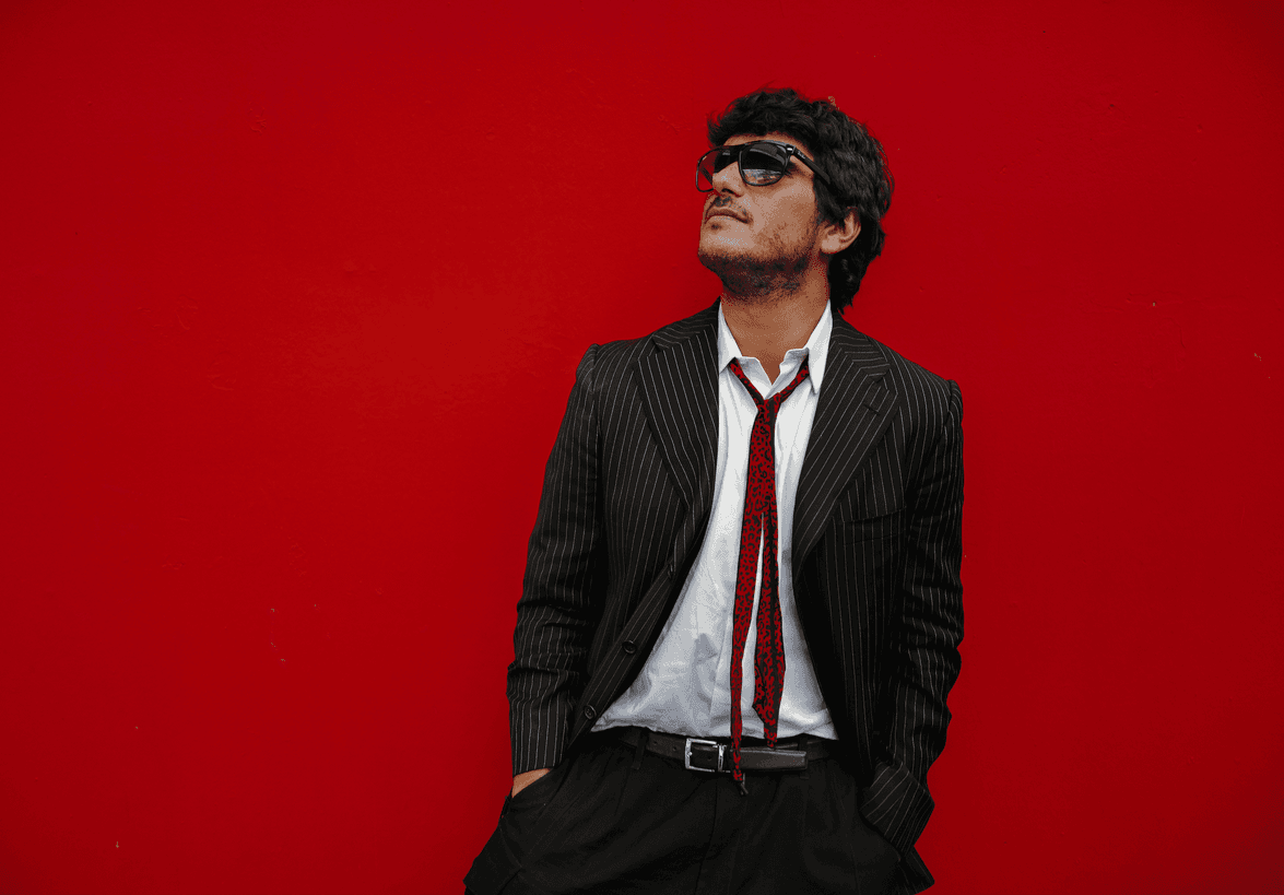 Sala Elassir de traje, corbata roja y lentes oscuros frente a una pared roja