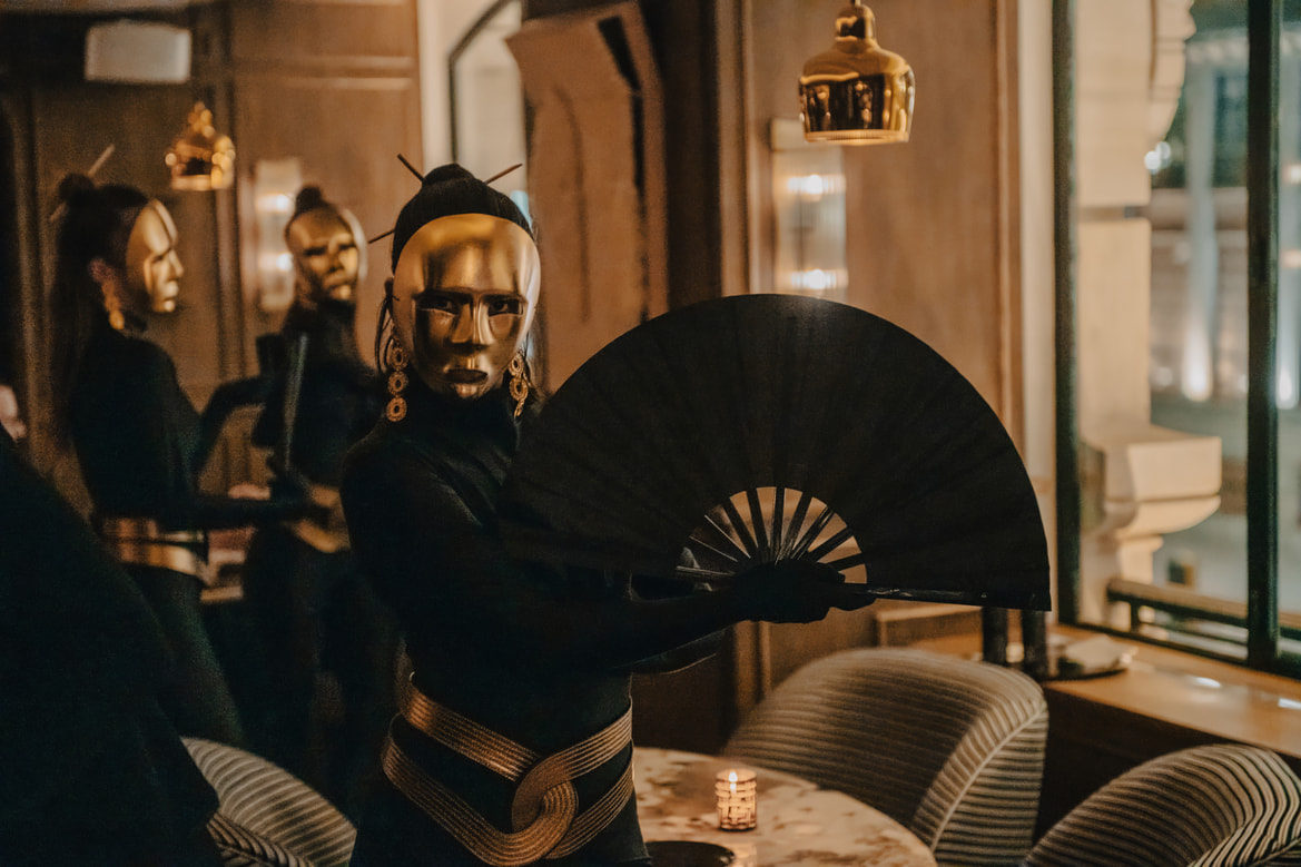 Artistas con máscaras doradas y ropa negra sosteniendo grandes abanicos en un bar