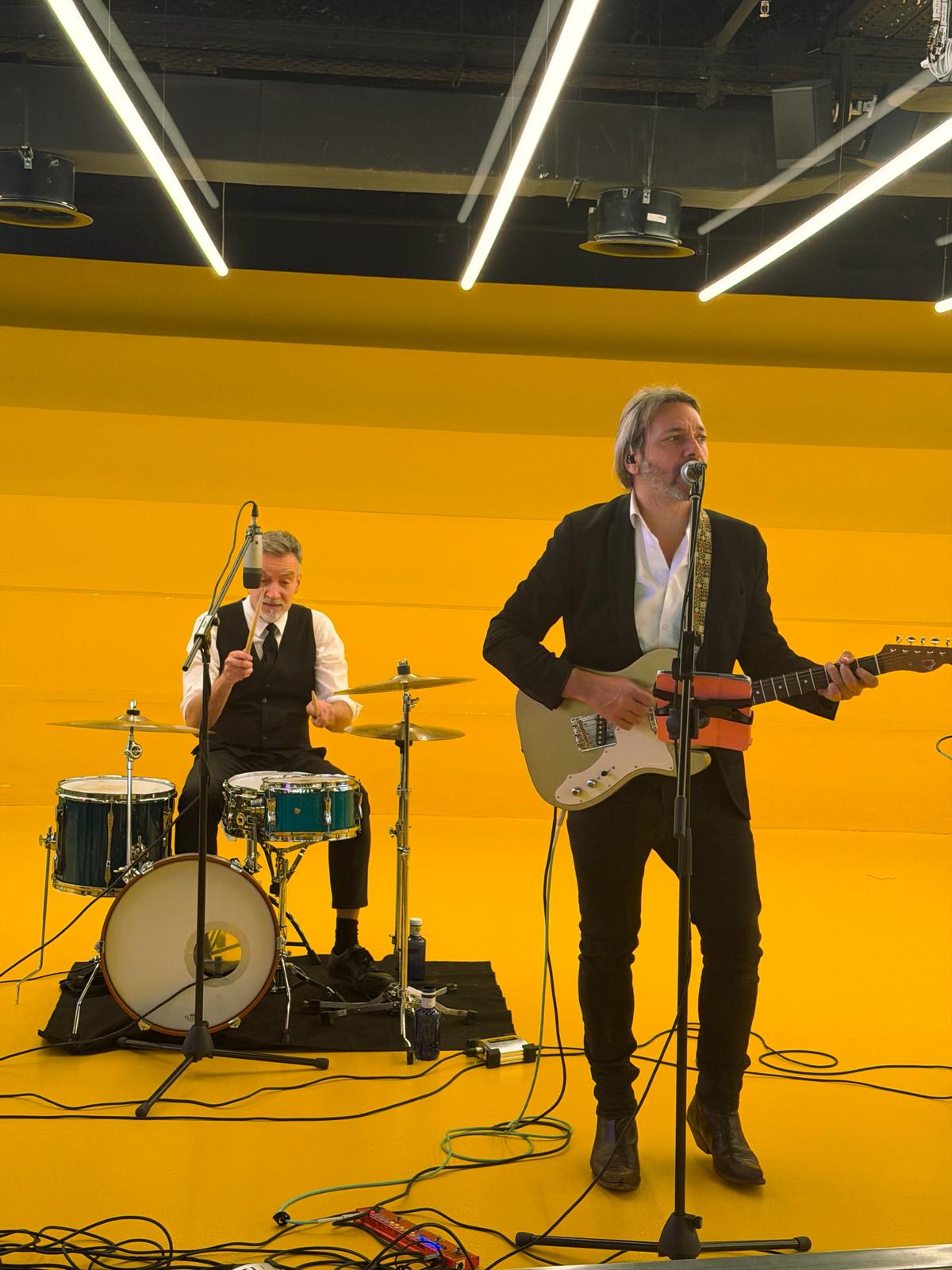 Cantante con guitarra eléctrica y baterista tocando en un escenario iluminado de amarillo