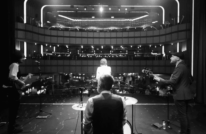 Foto en blanco y negro de una banda en el escenario de un teatro, vista desde atrás hacia las butacas