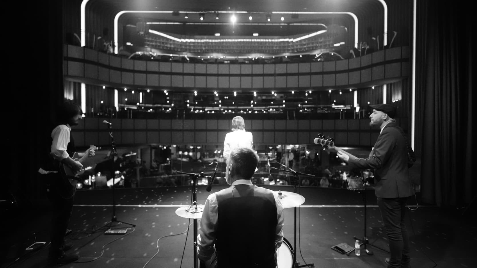 Foto en blanco y negro de una banda en el escenario de un teatro, vista desde atrás hacia las butacas