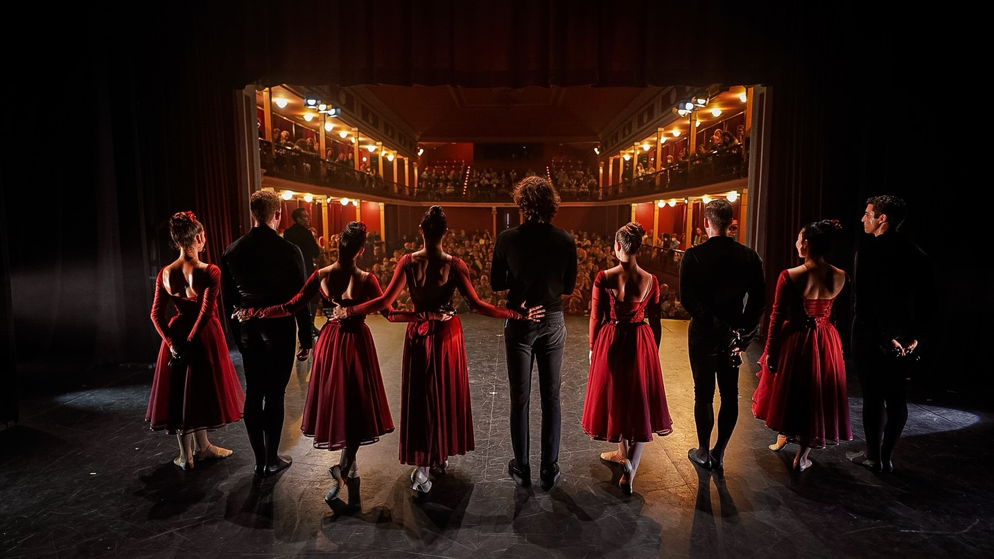 Bailarinas con vestido rojo y bailarines de negro tomados de la mano en el escenario, frente al público