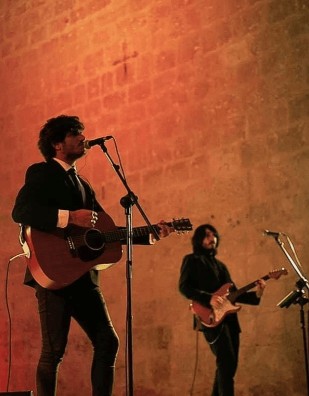Cantante con guitarra acústica y un guitarrista eléctrico tocando frente a un muro de piedra