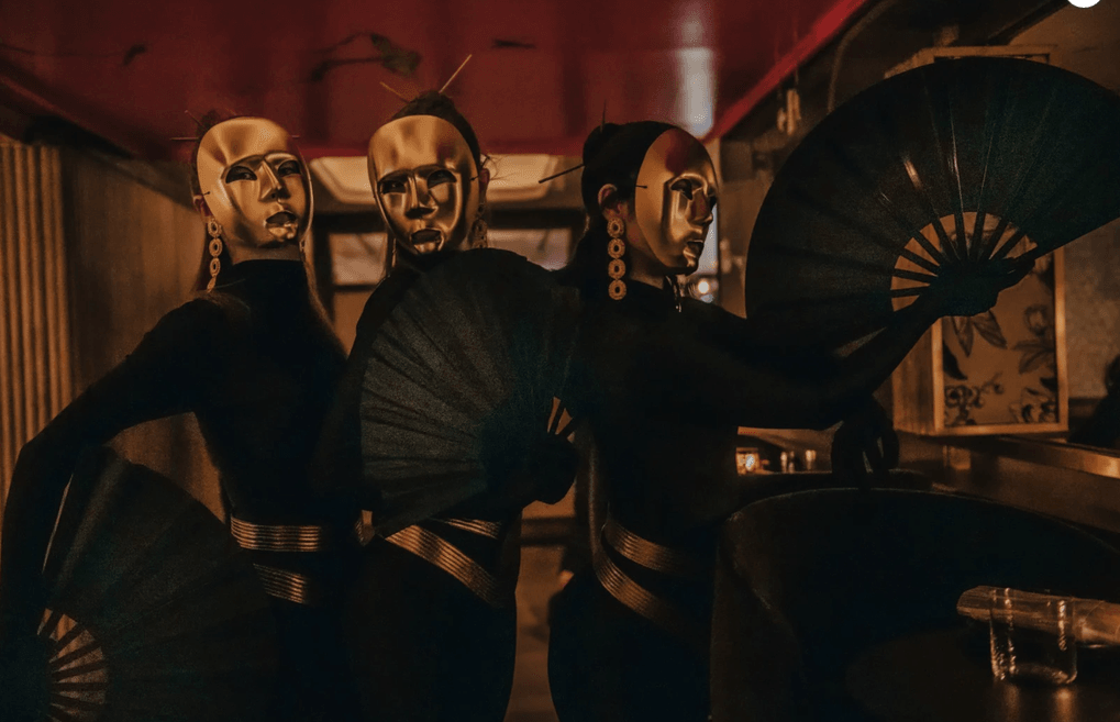 Tres artistas con máscaras doradas y ropa negra sosteniendo grandes abanicos